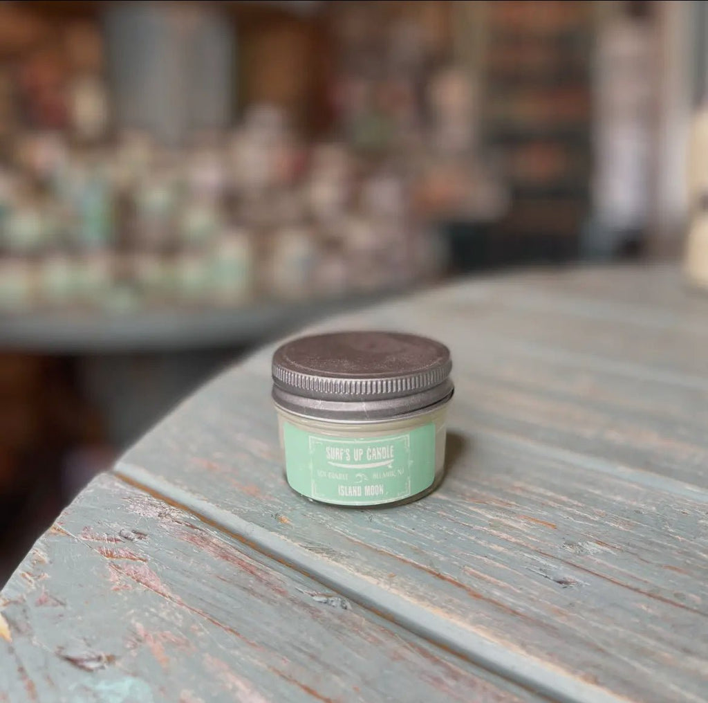 A small green jar labeled Surf’s Up - Island Moon Mason Jar Candle by Surfs Up Candles rests on a rustic wooden table. The softly blurred background evokes a cozy ambiance with hints of similar jars.