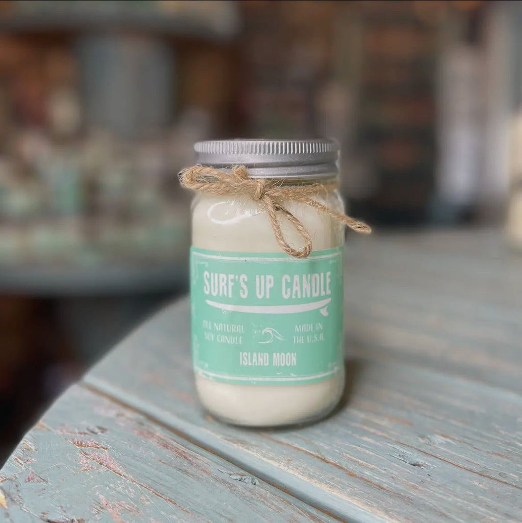 A Surfs Up Candle, branded as Surfs Up Candles, Island Moon soy candle with a light blue label sits on a distressed wood table. Made in the U.S.A., the reusable mason jar is elegantly tied with twine.