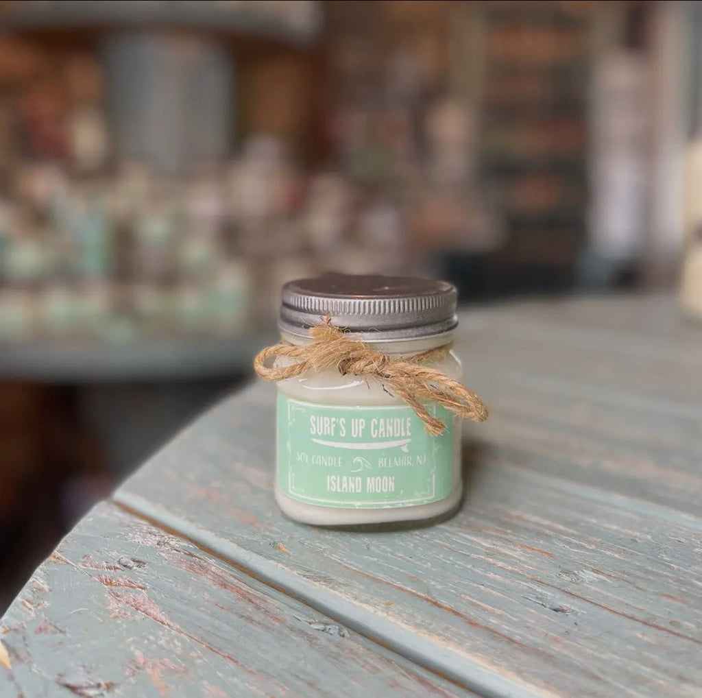 A small Surf’s Up - Island Moon Mason Jar Candle by Surfs Up Candles, made of natural wax with a light green hue and rustic twine bow, sits on a textured wooden table. Blurred shelves with more candles are in the background.