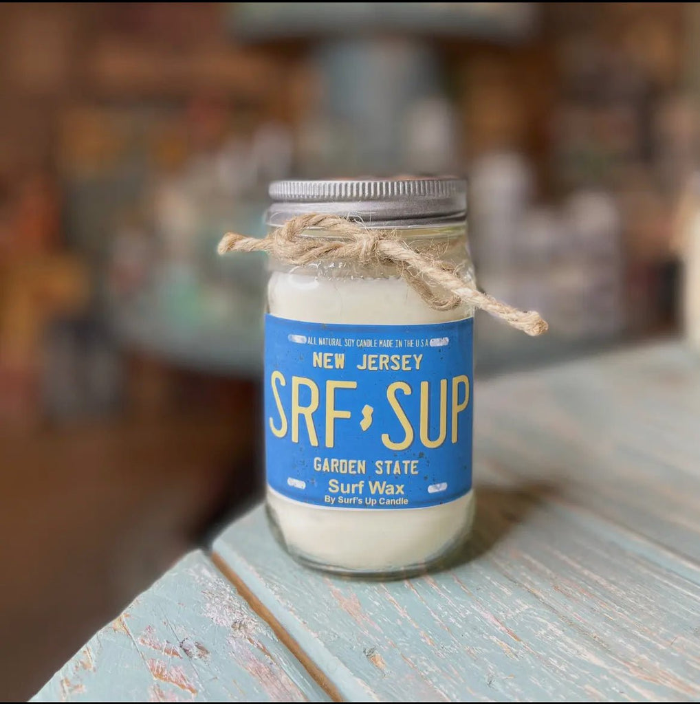 A glass jar candle named Surf’s Up - New Jersey License Plate Surf Wax Mason Jar Candle by Surfs Up Candles, made of all-natural soy wax with a twine bow on the lid, sits elegantly on a wooden surface against a blurred background.