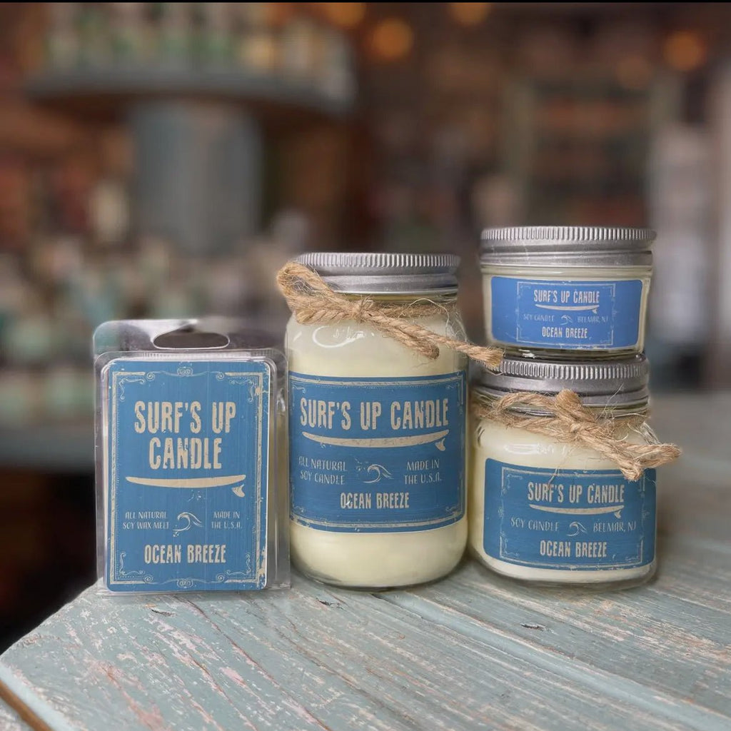 Four Surfs Up Candle products by Surf’s Up Candles on a wooden surface include three reusable Mason Jar candles with twine and one wax melt pack. All have blue labels with Ocean Breeze in white, containing soy wax and refreshing essential oils.