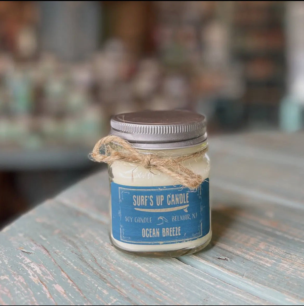 A small soy wax glass jar candle with a silver lid rests on a light blue wooden surface. The label reads Surf’s Up - Ocean Breeze Mason Jar Candle by Surfs Up Candles. A twine bow adorns the reusable Mason Jar, adding charm against a softly blurred background.