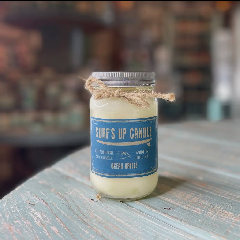 A weathered wooden table holds a Surfs Up Candles reusable Mason jar labeled Surf’s Up - Ocean Breeze. The pure soy wax candle features a rustic twine bow around its lid, exuding its essence amid a softly blurred background.