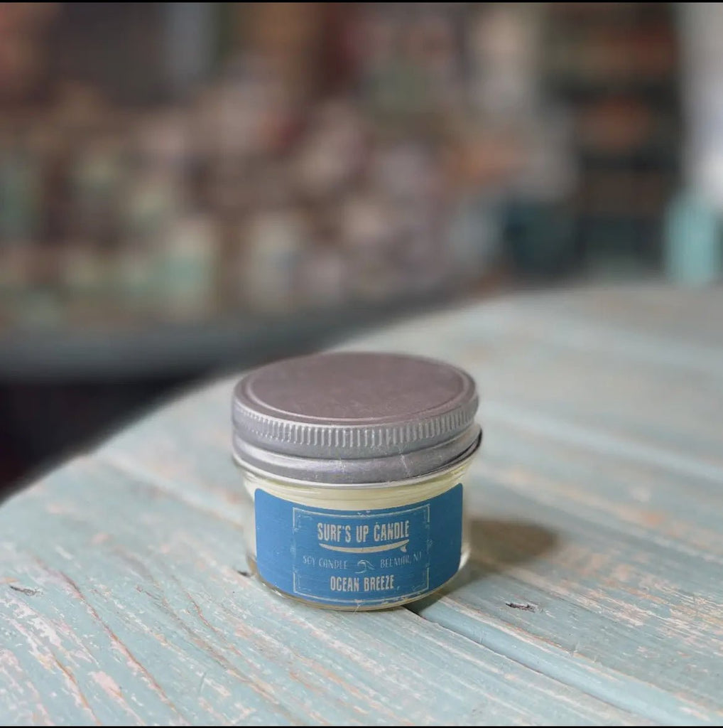 A small soy wax candle with a metal lid sits on a weathered, light blue wooden surface. The label reads Surf’s Up - Ocean Breeze Mason Jar Candle by Surfs Up Candles. The softly blurred background emphasizes the candle, infused with essential oils.