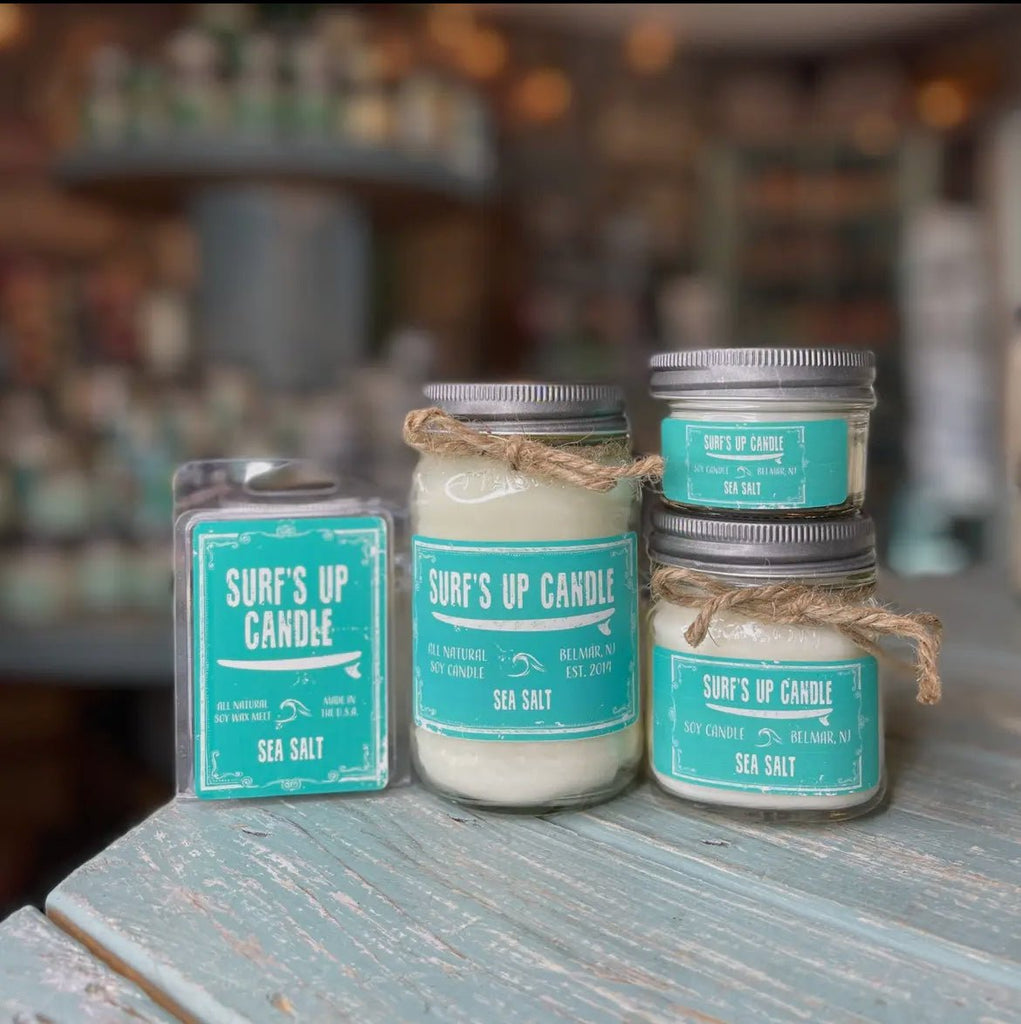 A Surfs Up Candle display presents sea salt-scented soy Mason Jar candles and a wax melt pack, all crafted from natural soy wax. Each item features a turquoise label with white text, showcased on wood against a backdrop of more candles.