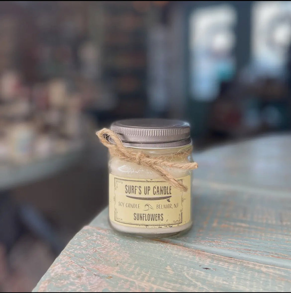 A Surf’s Up - Sunflowers Mason Jar Candle from Surfs Up Candles sits on a wooden table. A twine bow decorates the neck, and the soft-focus background highlights its floral fragrance.