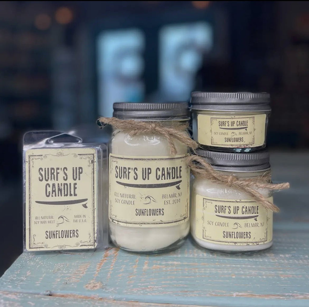 The image displays three Surfs Up Candles Surf’s Up - Sunflowers Mason Jar Candle products on a wooden table: two are small mason jars with silver lids, and one is a wax melt pack. Each has a rustic, off-white label tied with brown string, emitting a delightful floral fragrance.