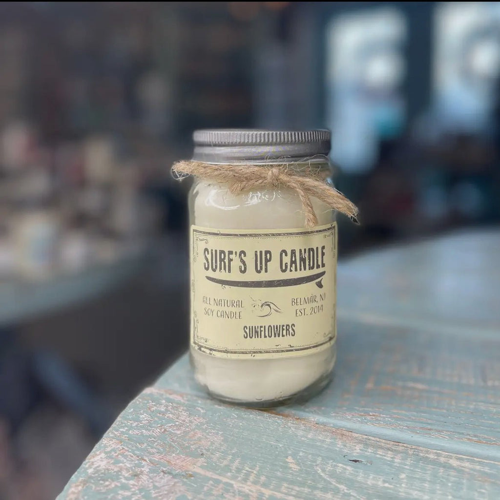 The Surfs Up - Sunflowers Mason Jar Candle by Surfs Up Candles features all-natural soy wax and a floral sunflower scent. It sits on rustic wood, topped with a metal lid and twine, against a softly blurred background that enhances its charm.