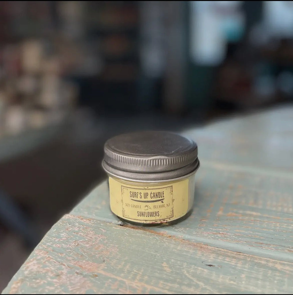 A small candle tin labeled Surfs Up - Sunflowers Mason Jar Candle by Surfs Up Candles sits on a wooden table with a metal lid. Made from soy wax, it emits a floral fragrance, while the softly blurred background emphasizes the candle.