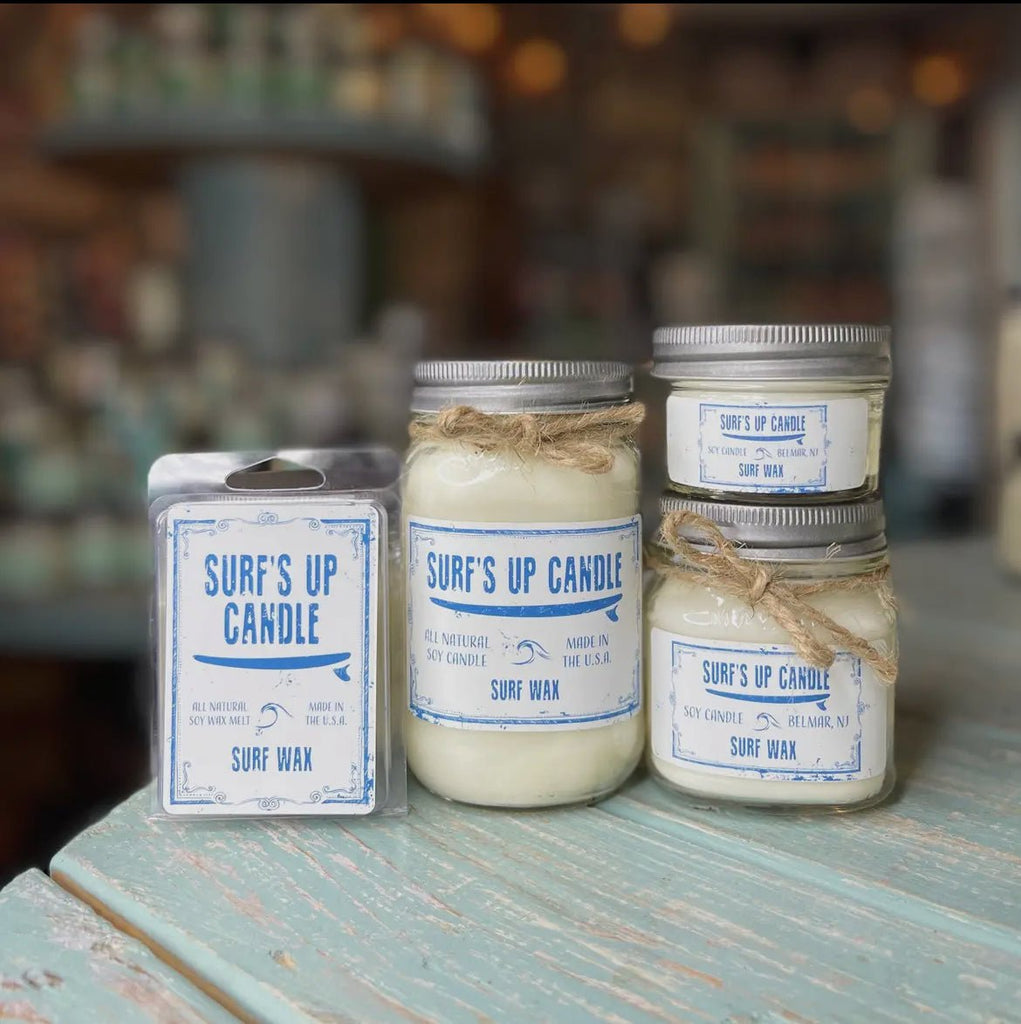 Four Surf’s Up Candles, three in reusable Mason jars with rope accents and one in a wax melt package, sit on a wooden table. These natural soy wax candles feature blue and white coastal-themed labels against a blurred store background, enhancing their charm.