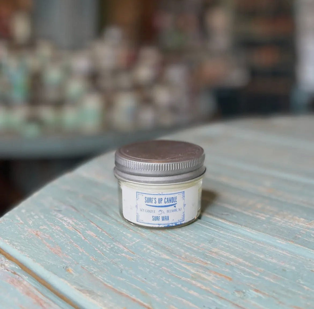 A small Surfs Up - Surf Wax Mason Jar Candle by Surfs Up Candles sits on a light blue wooden surface, crafted from natural soy wax. The background is blurred, showing more candles in reusable Mason Jars.