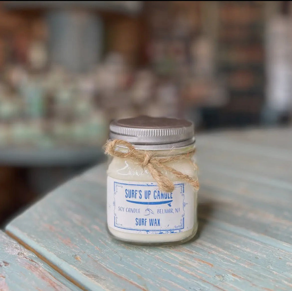 A Surfs Up - Surf Wax Mason Jar Candle by Surfs Up Candles, made of natural soy wax and tied with twine, rests on a light blue weathered wooden surface. The reusable Mason jar enhances the charm of the softly blurred rustic indoor setting.