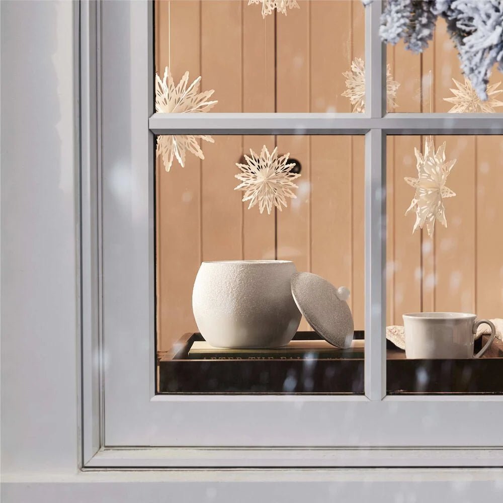 A cozy kitchen window features a white pot, cup, and paper snowflakes. The Thymes Fraiser Fir Flocked Novelty Statement Candle emits a warm glow as snow falls outside, enhancing the inviting wooden interior reminiscent of a fir forest.