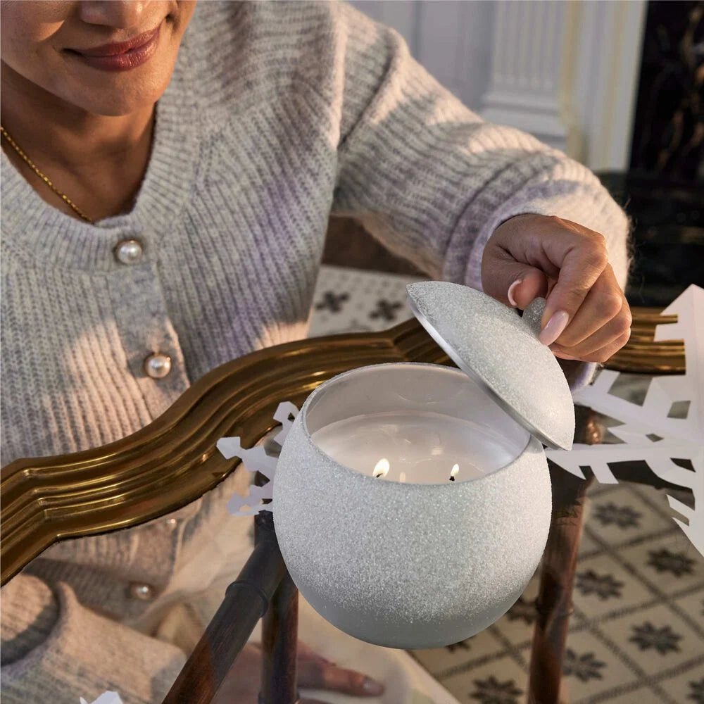 A cozy individual holds the lid of a Thymes - Fraiser Fir Flocked Novelty Statement Candle, featuring three lit wicks, while seated at a glass table with gold edges. A decorative paper snowflake completes the enchanting atmosphere.