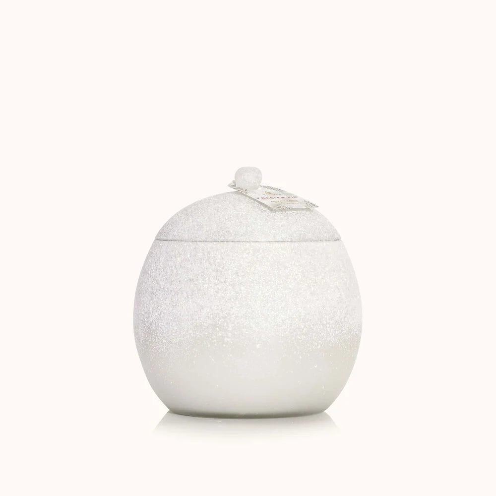 The Thymes Fraiser Fir Flocked Novelty Statement Candle, with its white round jar, fitted lid, and top knob, exudes elegance reminiscent of frosted glass decor. A textured surface and a tag on the lid enhance its charm against a plain light gray background.