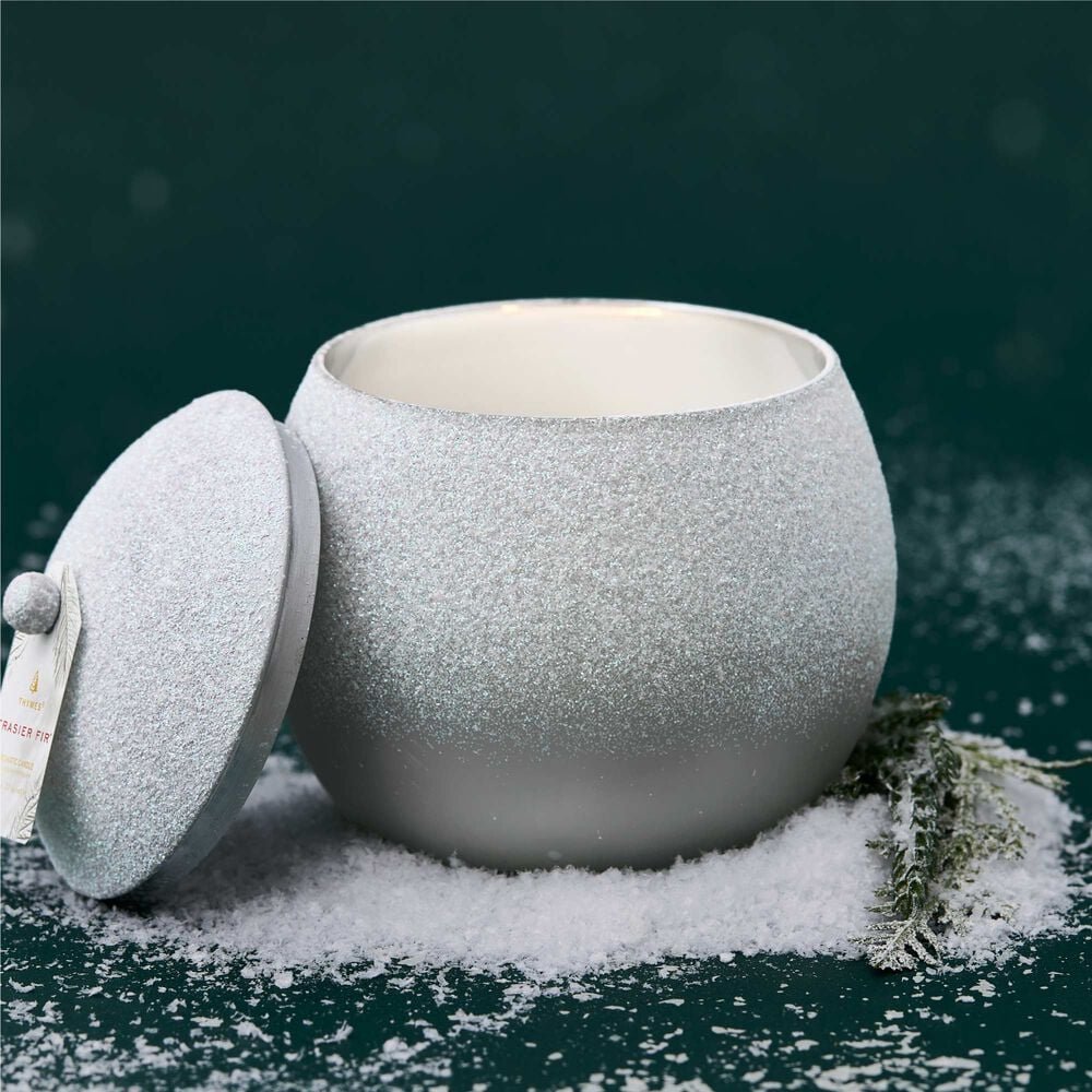 A Thymes - Fraiser Fir Flocked Novelty Statement Candle in a round, silver glittery jar without its lid sits amid white snow-like powder on a dark green surface, complemented by a sprig of greenery that captures the essence of Frasier Fir and enhances the winter-themed aesthetic.