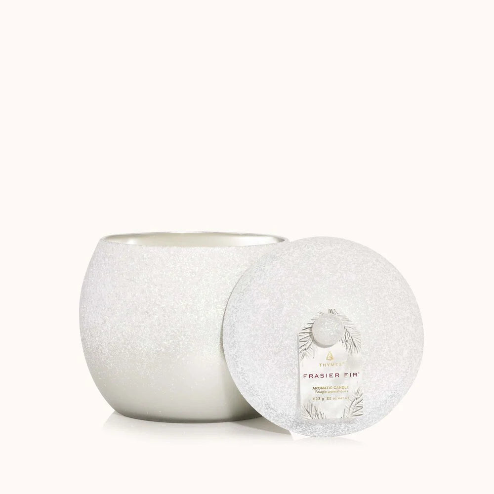 The Thymes - Fraiser Fir Flocked Novelty Statement Candle, from Thymes, features a smooth, rounded container with a matching lid adorned with pine illustrations. The lid rests alongside the candle on a light background, highlighting its elegant presence.