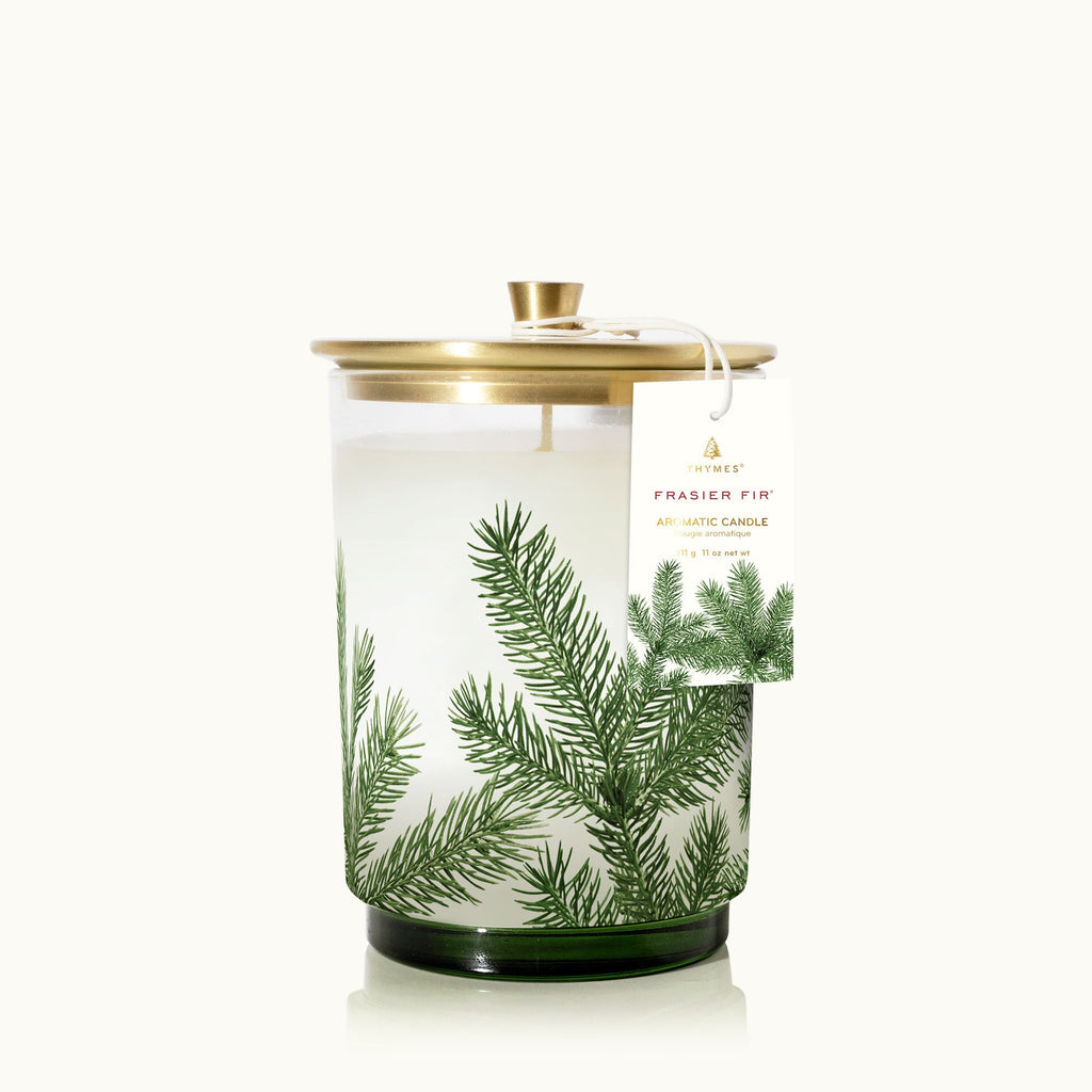 The Thymes Frasier Fir Medium Pine Needle Luminary 11.5oz Aromatic Candle features a white candle in a glass jar with green pine branches and a gold metal lid, complete with a festive tag—ideal for the holidays.