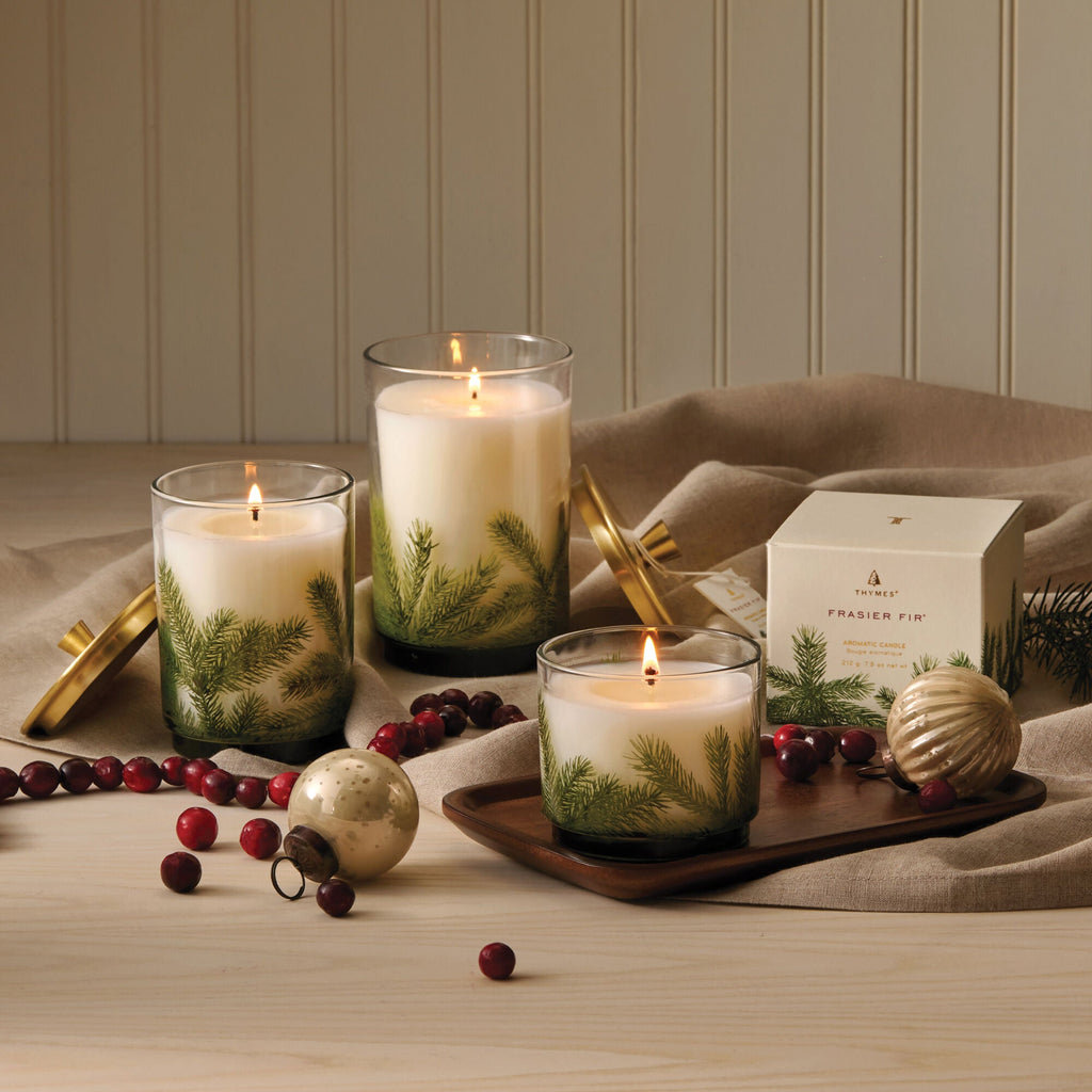 Three lit Thymes Frasier Fir Medium Pine Needle Luminary 11.5oz candles in glass holders with pine branch designs sit on a wooden surface, surrounded by cranberries, a gold ornament, a beige cloth, and the product box.