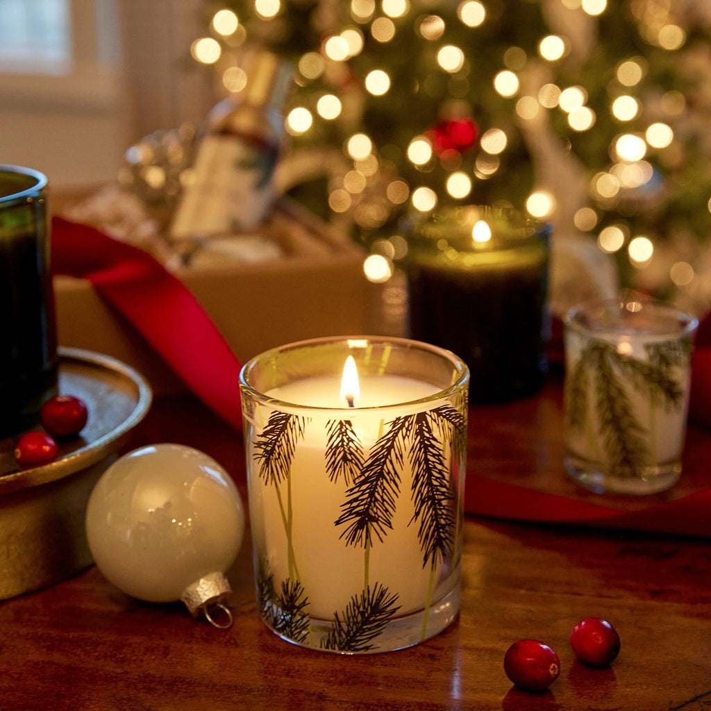 The Thymes - Frasier Fir Pine Needle 6.5oz Aromatic Candle, in a pine branch glass holder, sits on a wooden table with cranberries, a white ornament, ribbon, and a softly lit Christmas tree in the background.