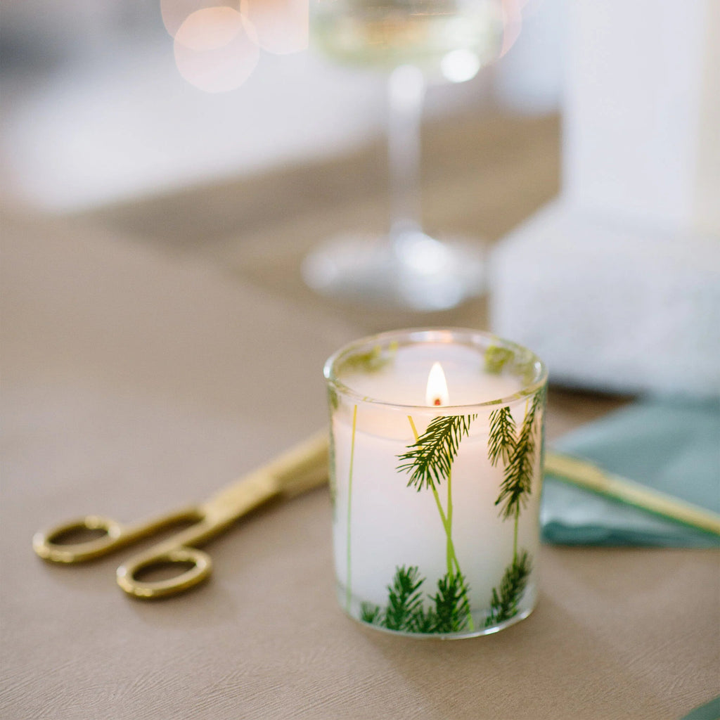 The Thymes Frasier Fir Pine Needle 6.5oz Aromatic Candle glows in its glass holder, surrounded by green pine branches, next to a gold wick trimmer and a folded teal napkin on a beige surface.