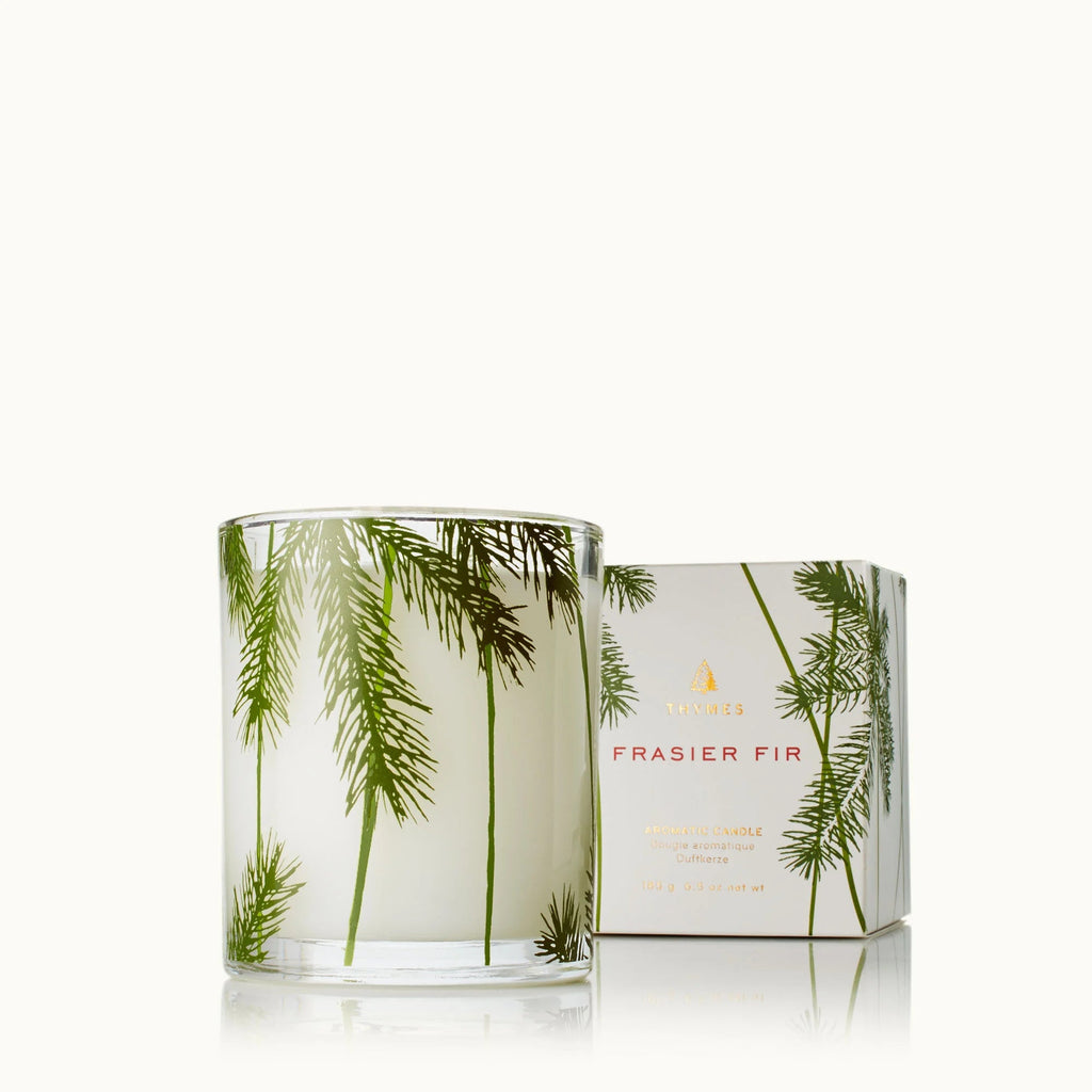 The Thymes Fraiser Fir Pine Needle 6.5oz Aromatic Candle, decorated with green fir branches, stands beside its matching box labeled in gold and red on a white background.