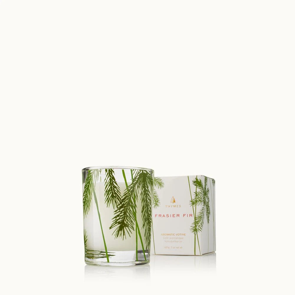 A Thymes - Fraiser Fir Pine Needle Votive 2oz Aromatic Candle decorated with green fir branches stands beside its matching box, both set on a plain white background. This candle offers the scent of fresh pine needles.