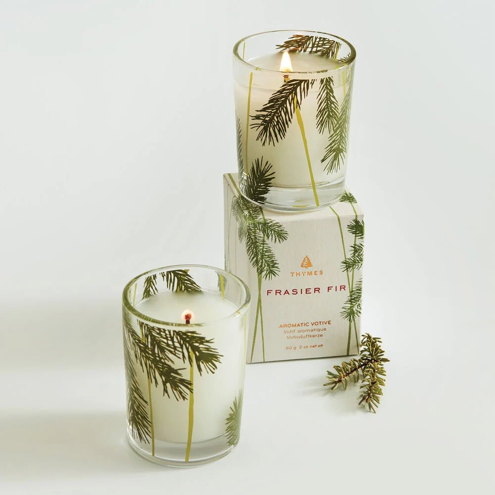 Two Thymes Frasier Fir Pine Needle 2oz votive candles, one lit and one unlit, are displayed with their box. A small pine branch rests beside these aromatic holiday candles by Thymes.