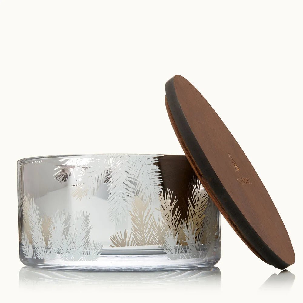 The Thymes - Frasier Fir Silver 40oz Aromatic Candle features white pine branch designs on clear glass with a round dark wooden lid, making it a luxurious centerpiece from Thymes that stands out against a simple, light background.
