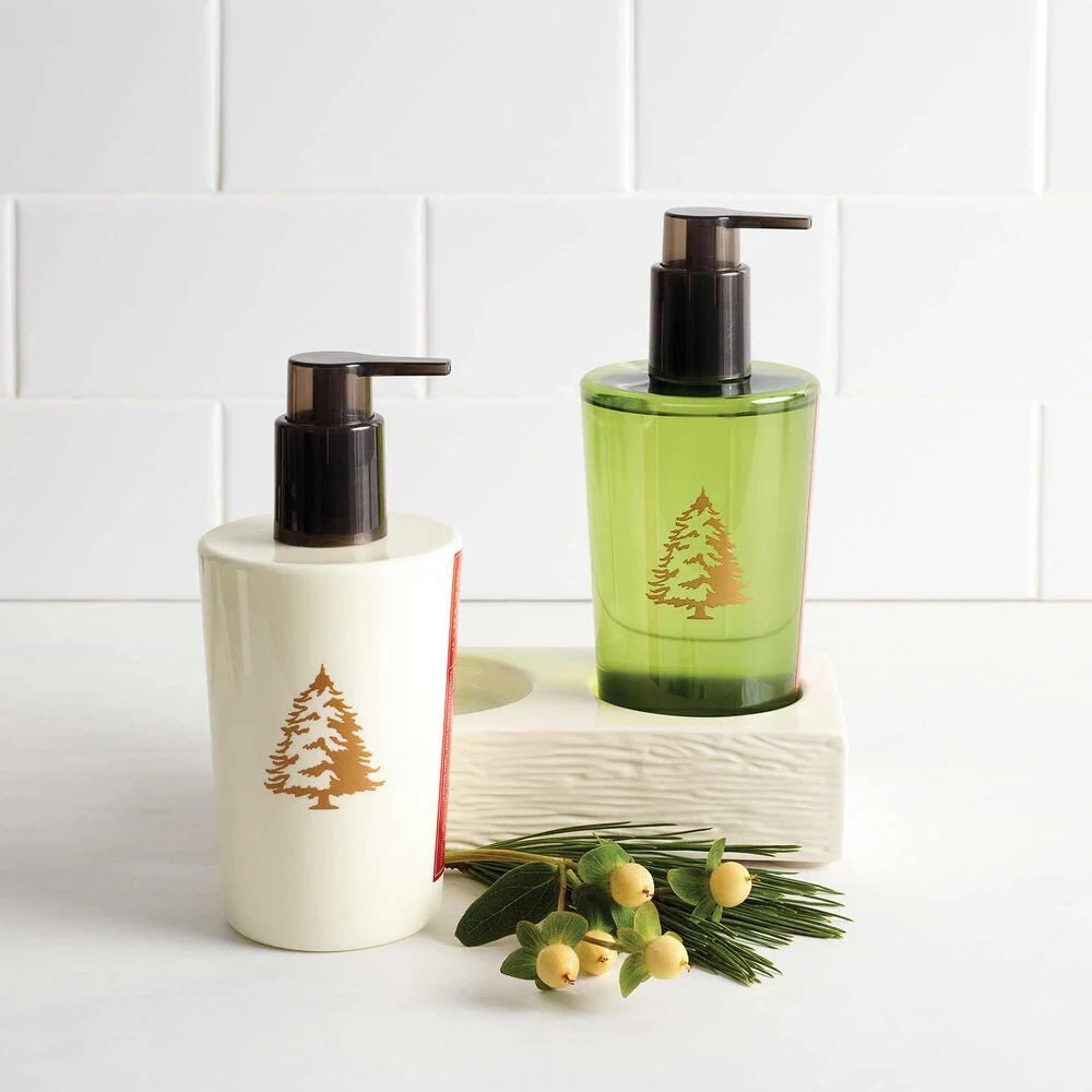 The Thymes Frasier Fir Sink Set features two soap dispensers, in white and green with a pine tree design, set on a log-like holder. A pine needle and white berry sprig complements the setup against a tiled backdrop, ideal for hand wash or lotion.