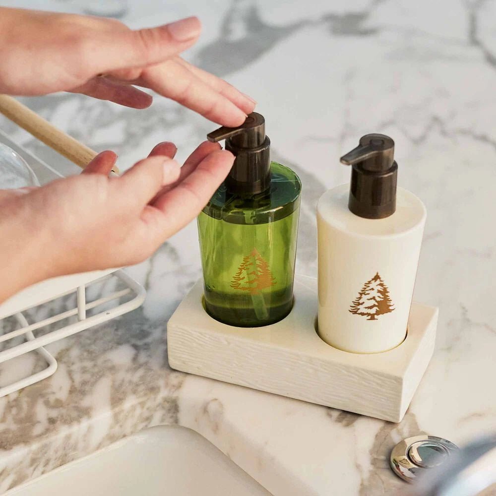 Hands pump Thymes - Frasier Fir hand wash from green and white bottles adorned with a tree design on a bathroom counter. Both bottles, in a wooden tray, complement the nearby white dish rack and chrome sink, enhancing the serene ambiance.