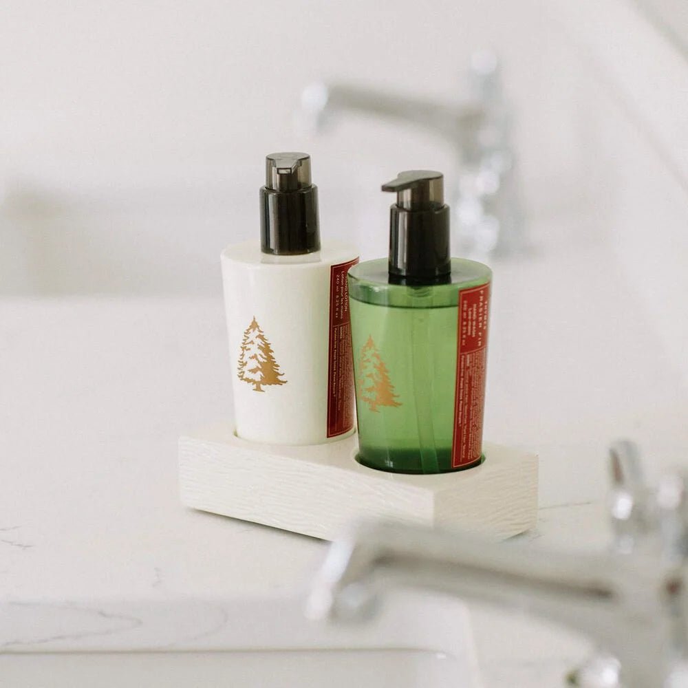 The Thymes Fraiser Fir Sink Set, featuring cream and green pump bottles with tree illustrations for hand wash and lotion, rests elegantly on a white tray atop a marble countertop with a silver faucet, adding a touch of nature to the bathroom.