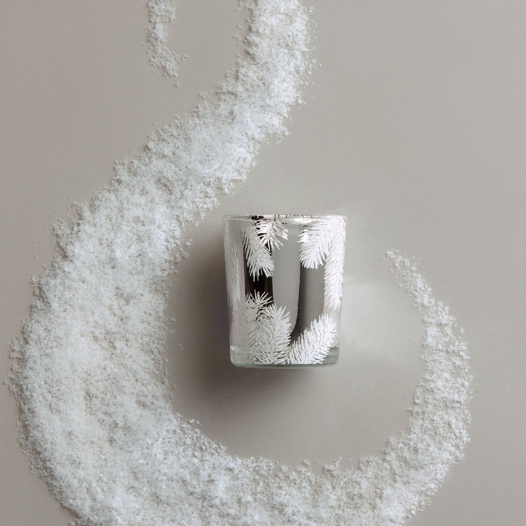 The Thymes - Frasier Fir Statement Boxed Votive 2oz Aromatic Candle, adorned with white pine and Siberian Fir motifs, rests on a light gray surface, encircled by a swirling trail of artificial snow.