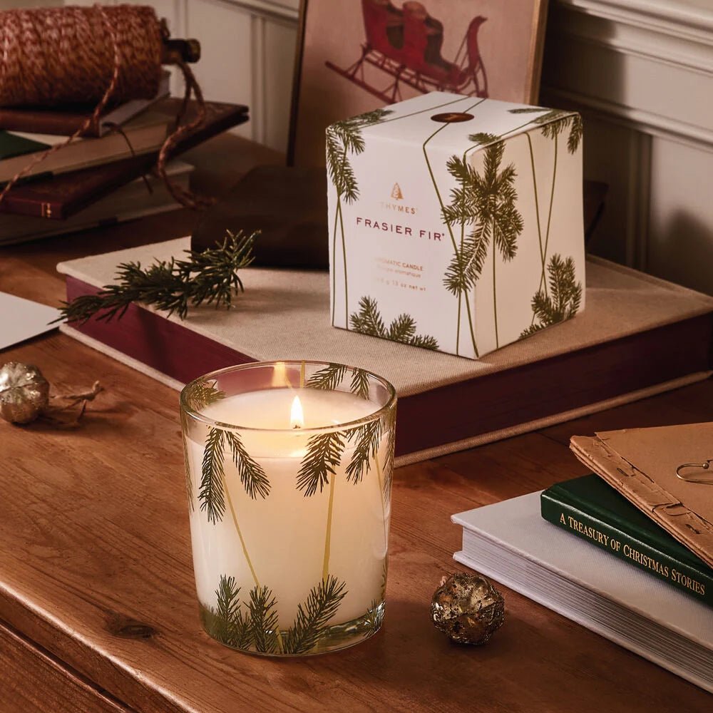 The Thymes - Frasier Fir 13oz Pine Needle Poured Candle in a glass holder with pine branch accents rests on a wooden desk beside books, a gold ornament, and its matching boxed candle, creating a warm holiday ambiance.