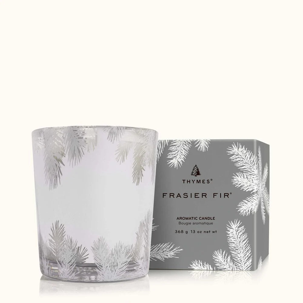 The Thymes Frasier Fir 13oz Statement Poured Candle sits in glass with a silver pine needle design, next to a matching grey box. Both display elegant white pine needle illustrations and are branded Thymes.