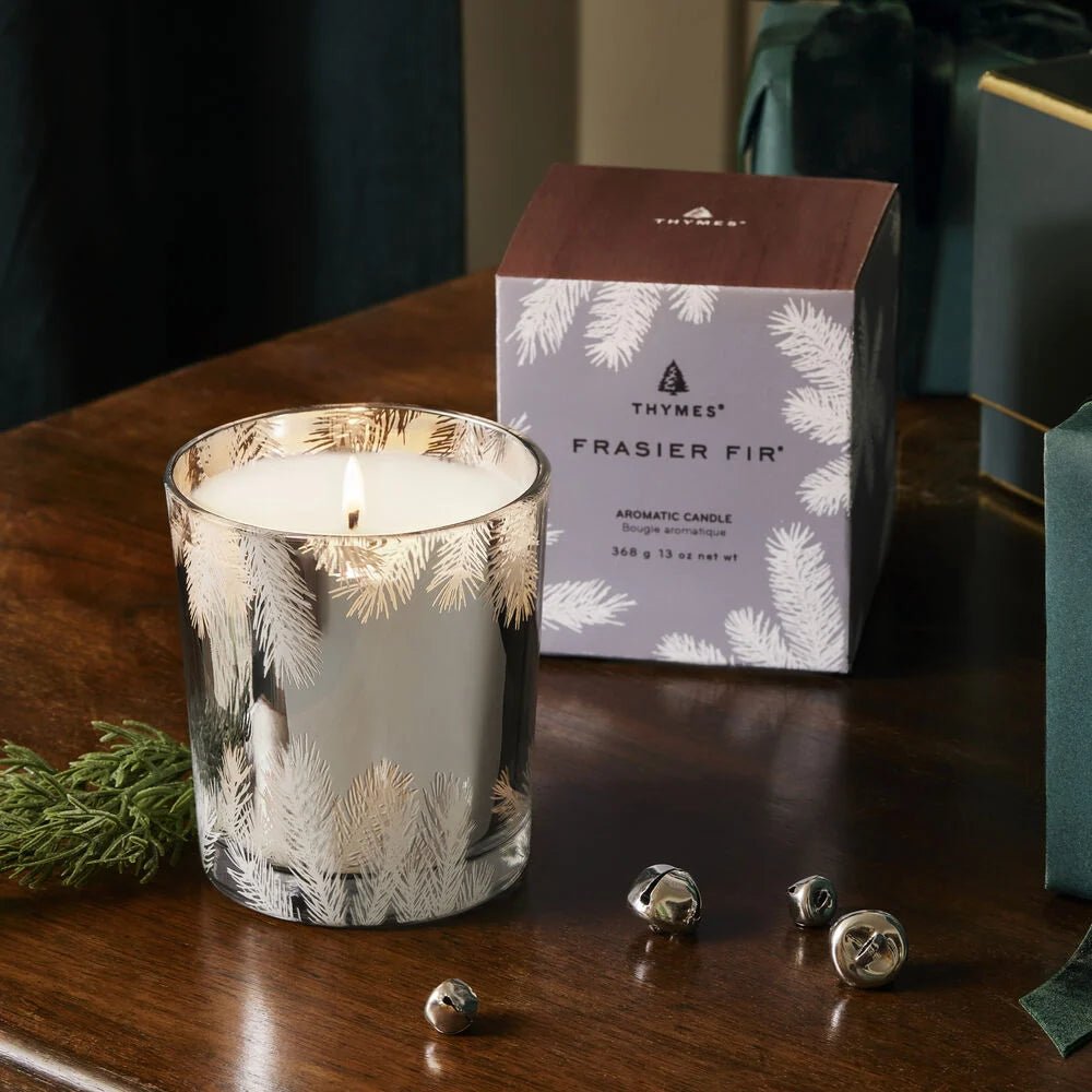 The Thymes - Frasier Fir 13oz Statement Poured Candle, in a glass holder with a pine needle design, sits beside its box on a table decorated with greenery, jingle bells, and a wrapped gift.