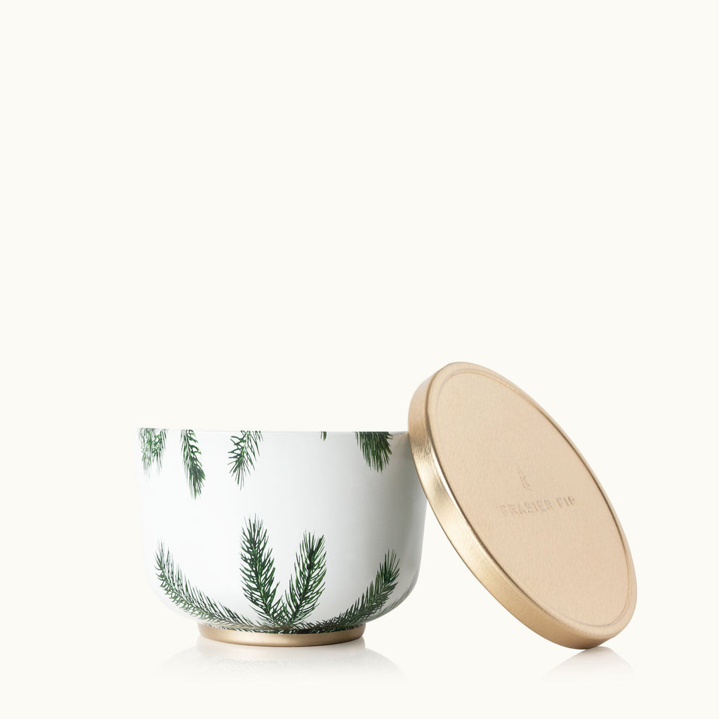 The Thymes - Frasier Fir 6.5oz Candle Tin features green pine tree illustrations and a separate circular gold lid embossed with Frasier Fir, all against a light background.