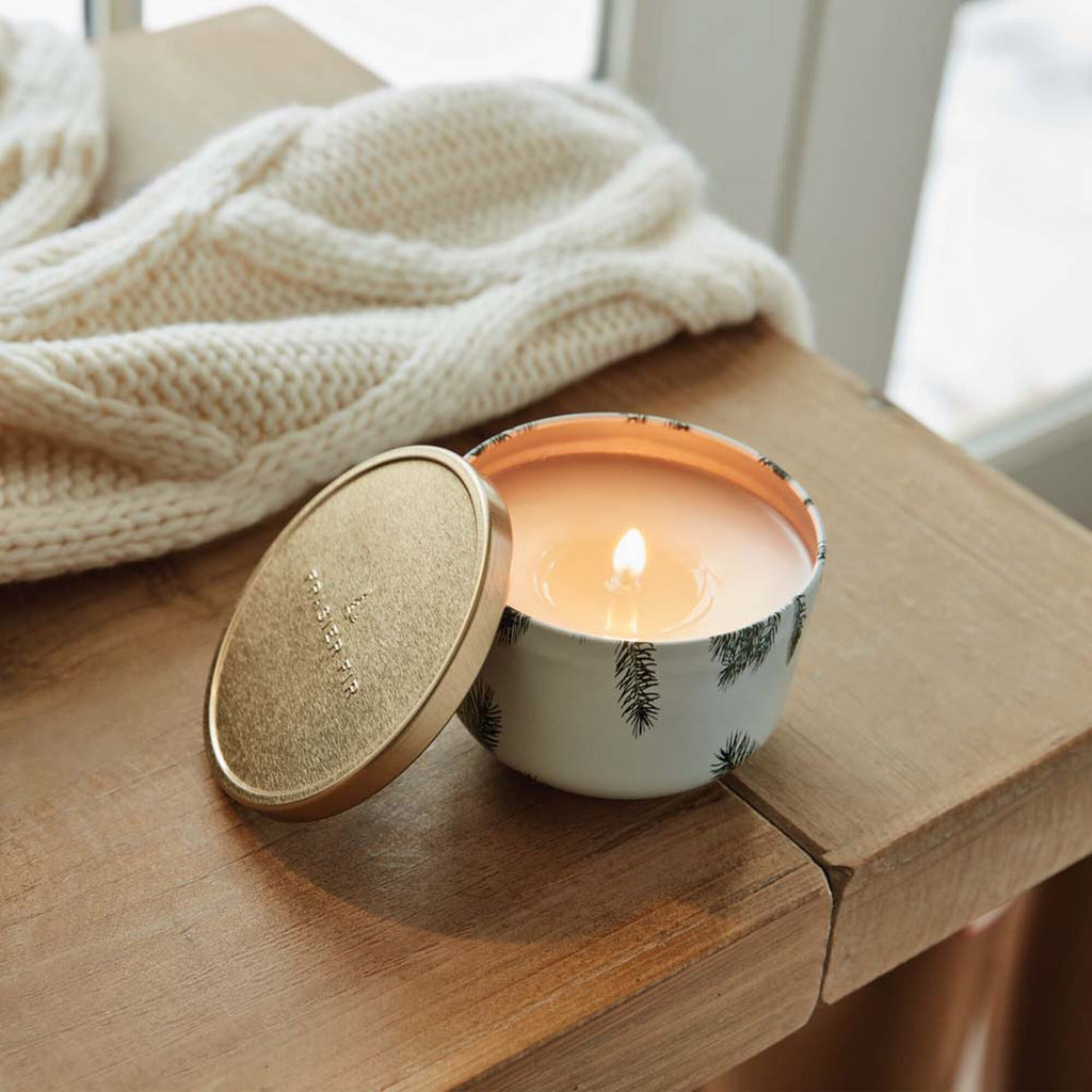 The Thymes - Frasier Fir 6.5oz Candle Tin with Gold Lid sits on a wooden table, its pine tree patterns beautifully complementing the setting. Its scent gently fills the room as a cream-colored knitted blanket drapes near a window in the background.