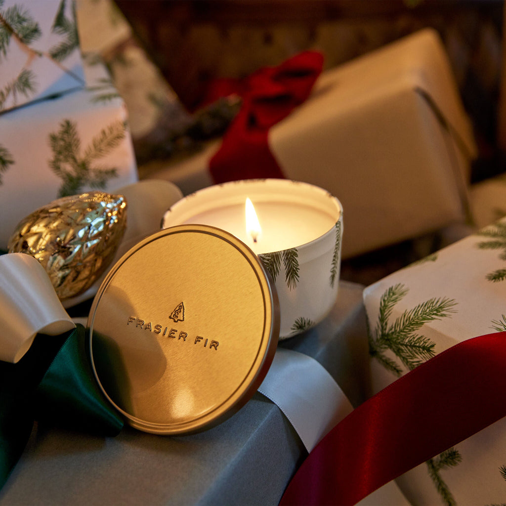 A Thymes Frasier Fir 6.5oz Candle Tin with Gold Lid flickers among gifts wrapped in festive pine branch paper, with colorful ribbons and an ornate gold pinecone decoration, creating a cozy holiday atmosphere.