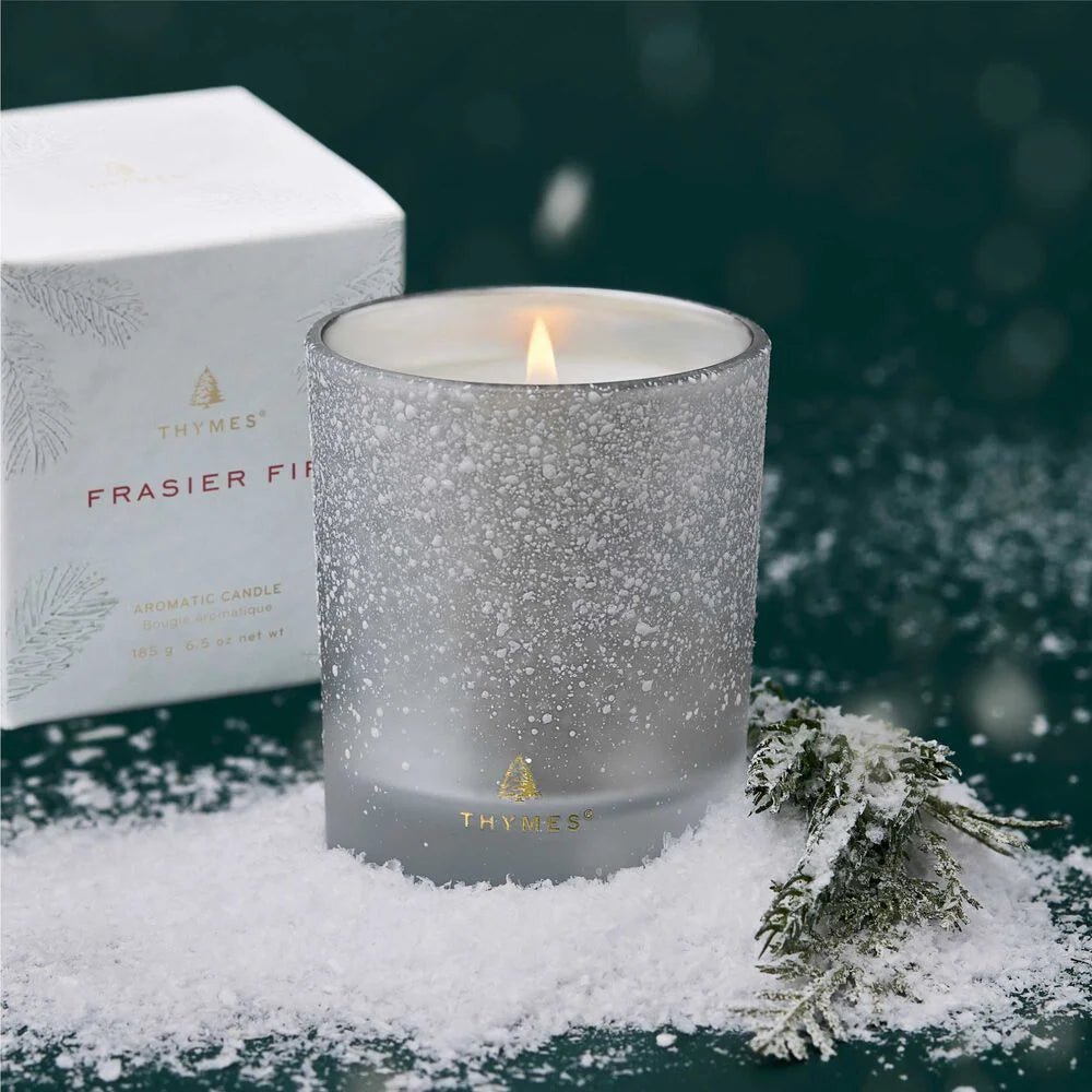 The Thymes - Frasier Fir Flocked 6.5 oz Poured Candle in a frosted glass holder fills the air with fresh Siberian Fir scent, displayed on artificial snow with a pine branch and its white box, set against a dark green background.
