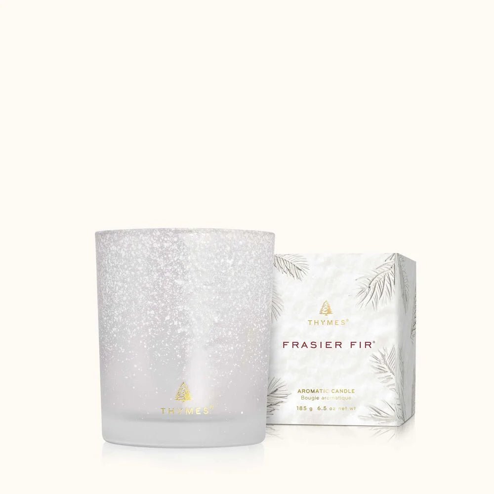 A white, speckled Thymes Frasier Fir Flocked 6.5 oz Poured Candle sits next to a matching box labeled “Thymes Frasier Fir Aromatic Candle” with silver pine needle designs on a white background.