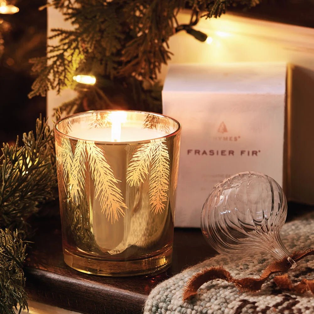 The Thymes Frasier Fir Gilded 13 oz Poured Candle, in a gold fir-branch glass holder, sits beside its white box, an ornament, and pine branches for a festive holiday display.