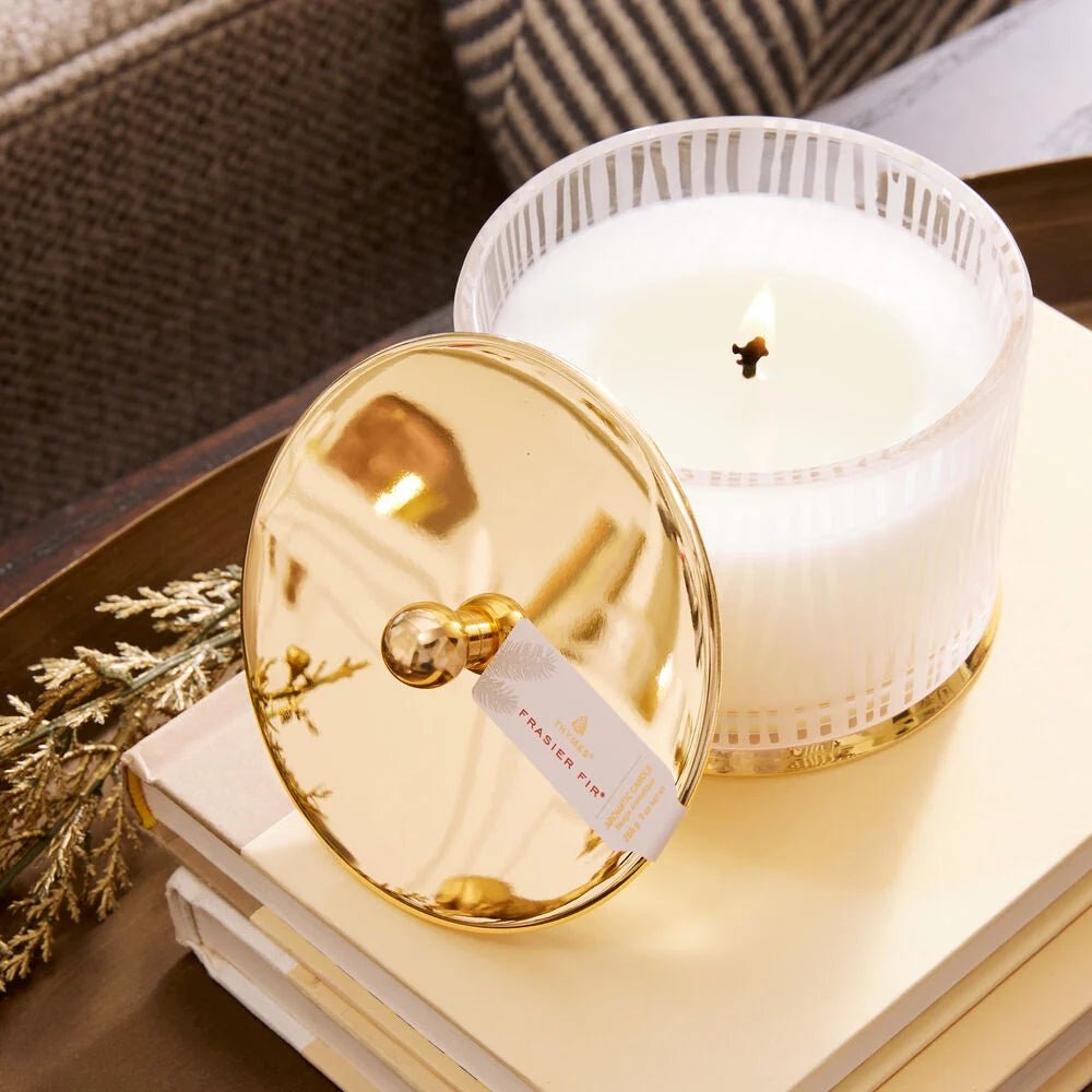 The Thymes Frasier Fir Gilded Frosted Wood Grain Candle in a ribbed glass jar with a gold lid sits atop cream books next to dried foliage, displayed on a brown tray.