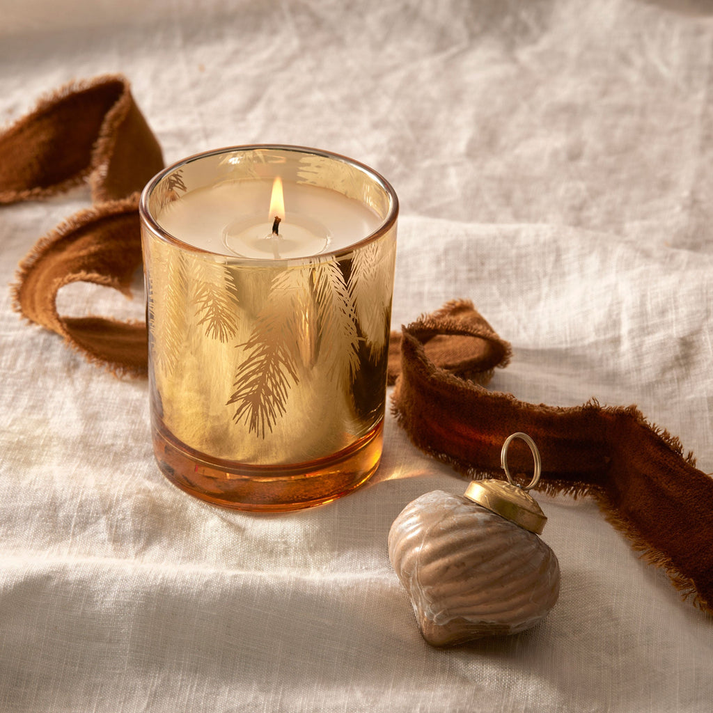 The Thymes Frasier Fir Gilded Gold Poured Candle, housed in a gilded glass holder with golden leaf patterns, sits on a cream cloth. Beside it lies a brown ribbon and a small metallic heart-shaped ornament, creating a warm and decorative scene.