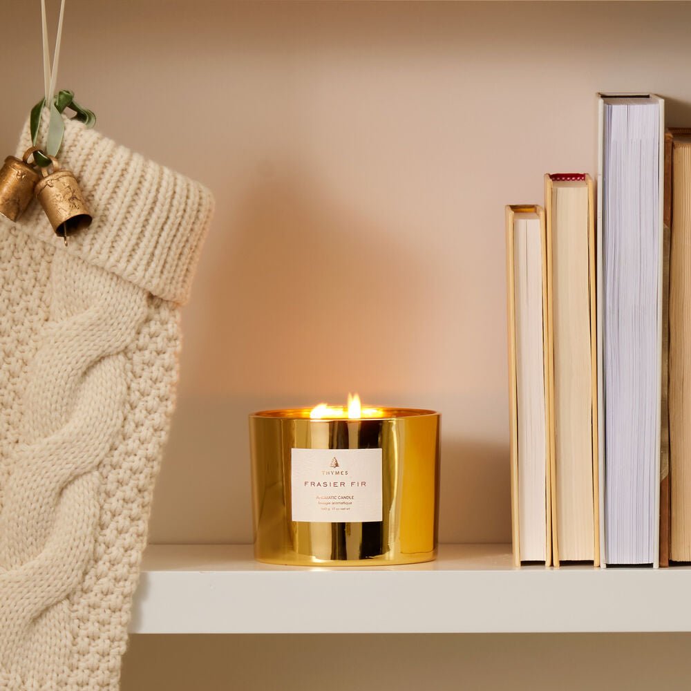 The Thymes Frasier Fir Gold 3-Wick Candle by Thymes burns on a white shelf beside closed books and a cream knit Christmas stocking adorned with small gold bells.