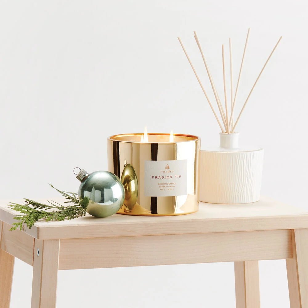 The Thymes - Frasier Fir Gold 3-Wick Candle glows on a light wood table with a green ornament, pine branch, and white diffuser nearby, filling the room with Thymes’ signature mountain fresh fragrance.