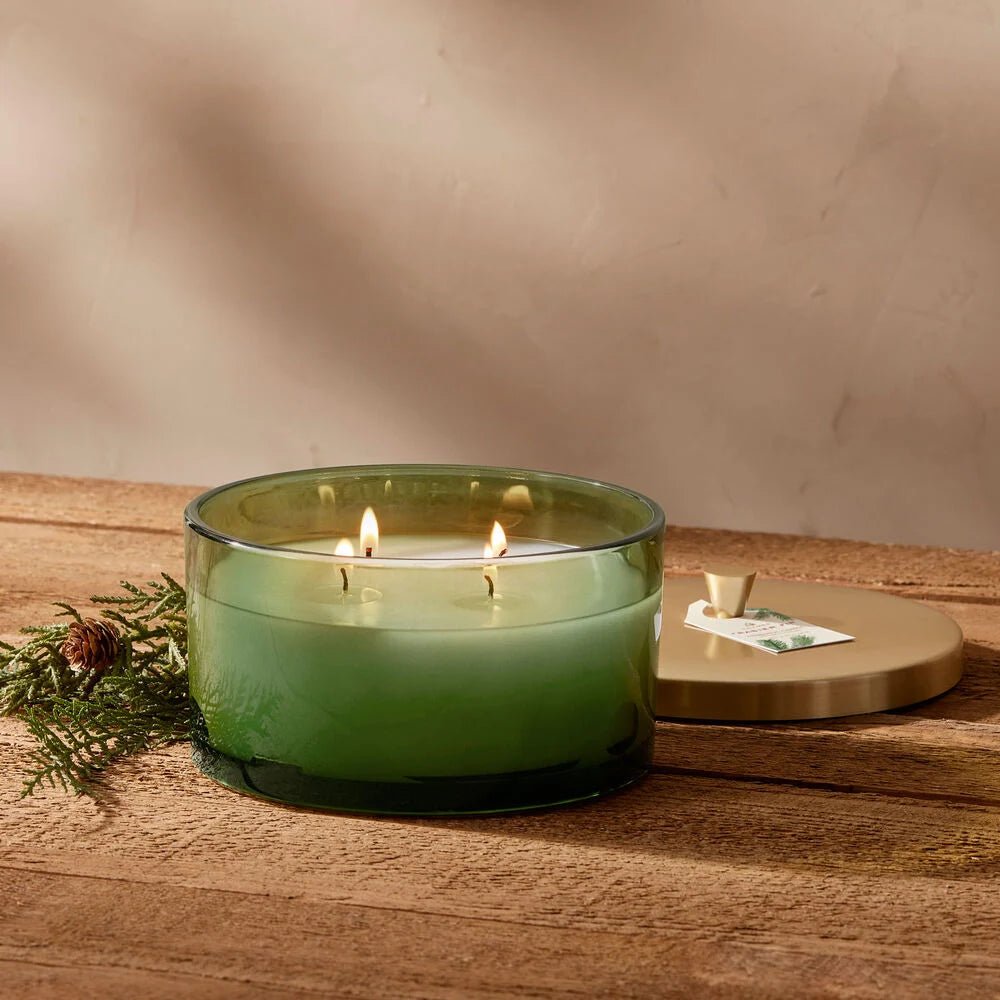 The Thymes Frasier Fir Green 4-Wick Candle sits on a wooden surface, its gold lid nearby. A pinecone with evergreen rests beside it, enhancing the Siberian fir fragrance that fills the air.