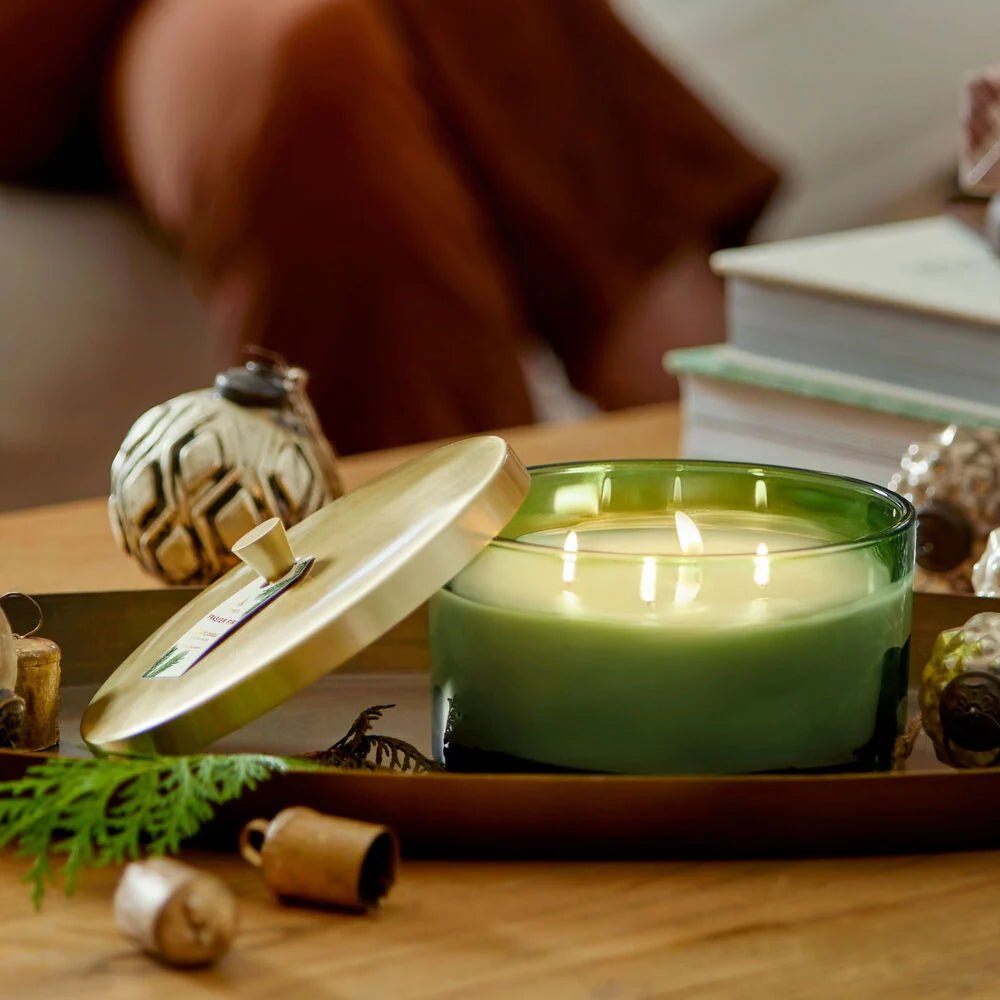 A Thymes Frasier Fir Green 4-Wick Candle glows on a rectangular tray with its gold lid beside it, surrounded by decor pieces; a book rests in the background atop a wooden table.