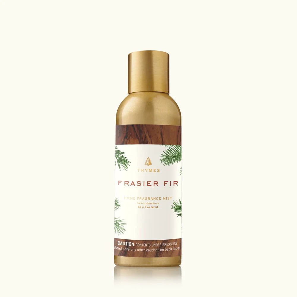 A gold bottle of Thymes Frasier Fir Home Fragrance Mist 3oz serves as an elegant air freshener, featuring green fir branches with white text on the label. Gold cap and accents add to its charm, and a warning at the bottom notes contents are under pressure.