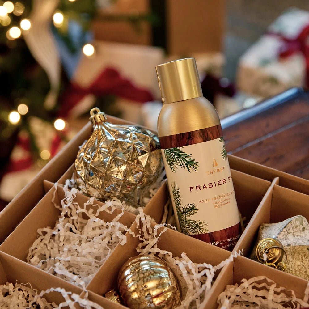 A box with shredded paper holds a Thymes - Frasier Fir Home Fragrance Mist 3oz and decorative ornaments: a silver geometric sphere and a gold ribbed sphere. In the blurred background, a Christmas tree stands tall, surrounded by beautifully wrapped gifts.